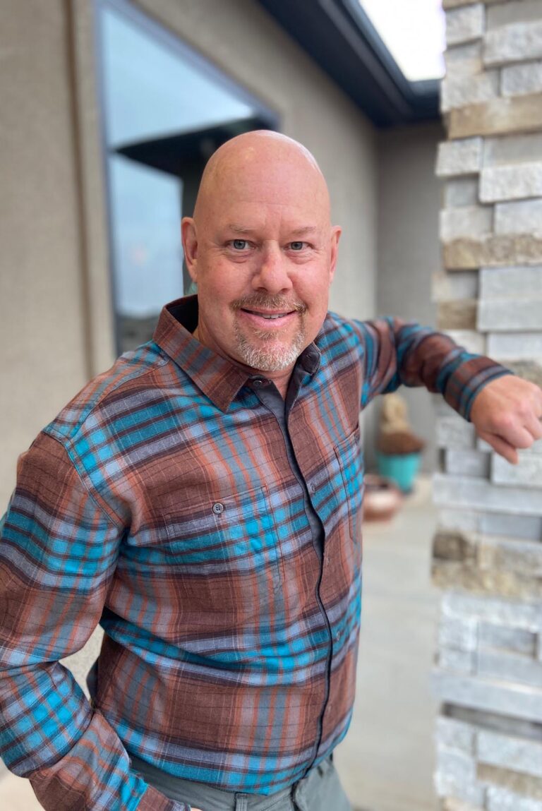 A man in a plaid shirt with blue and brown stripes, standing beside a stone pillar outdoors. The background shows part of a building and open space.