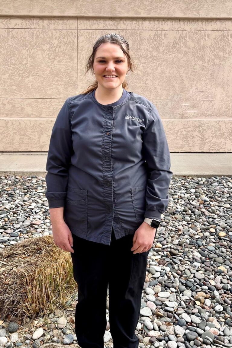 A woman standing outdoors on a surface with rocks and dry grass, wearing a dark gray jacket with a logo on the chest, black pants, and a smartwatch. Background includes a beige wall.