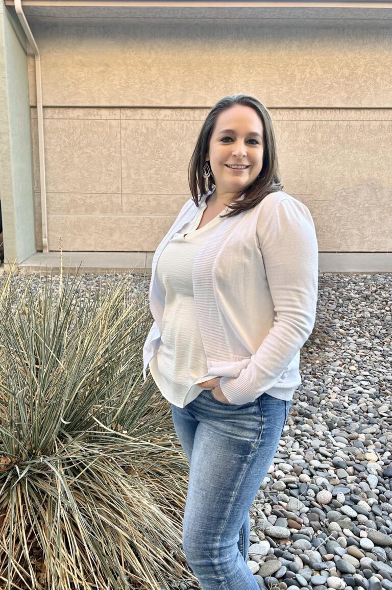 A woman standing outdoors on a surface with small rocks, wearing a white cardigan over a white top and blue jeans, with hands in pockets. A plant with thin leaves is on the left; the background shows a beige wall.