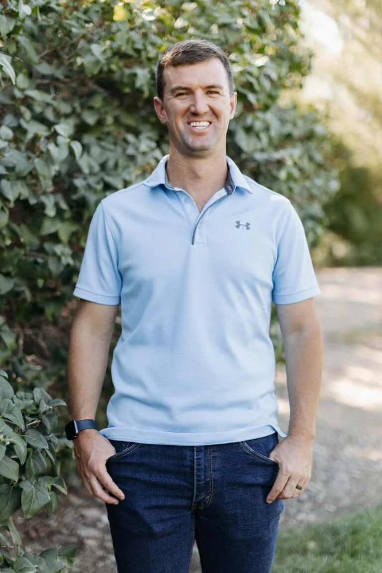 Dr. Ryan is standing outdoors in front of green foliage, wearing a light blue Under Armour polo shirt, dark blue jeans, a black smartwatch on the left wrist, and a ring on the left hand.
