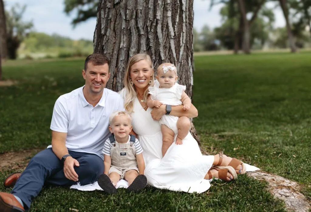 The image shows a family of four sitting on the grass in front of a large tree. The adults are wearing white outfits, and the children are dressed in a white headband and overalls. They are enjoying a moment together outdoors with a grassy area, trees, and a cloudy sky in the background.