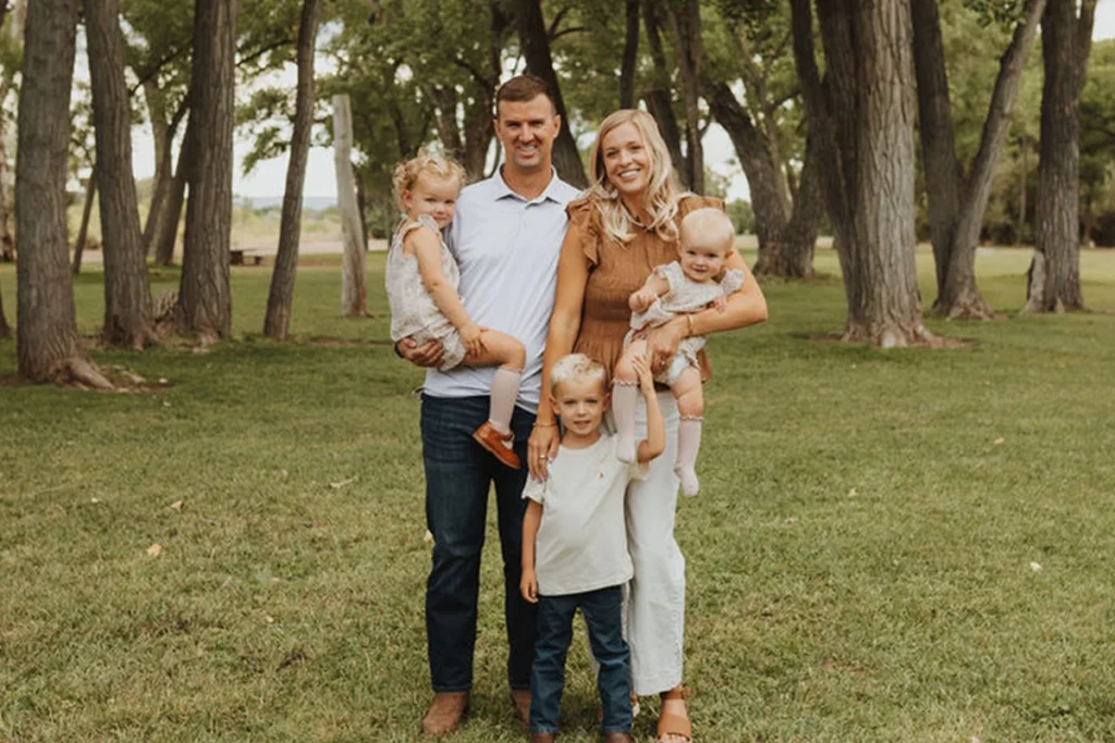 A family picture taken outdoors on grassy area with tall trees, two adults holding small children, and one child standing in front.