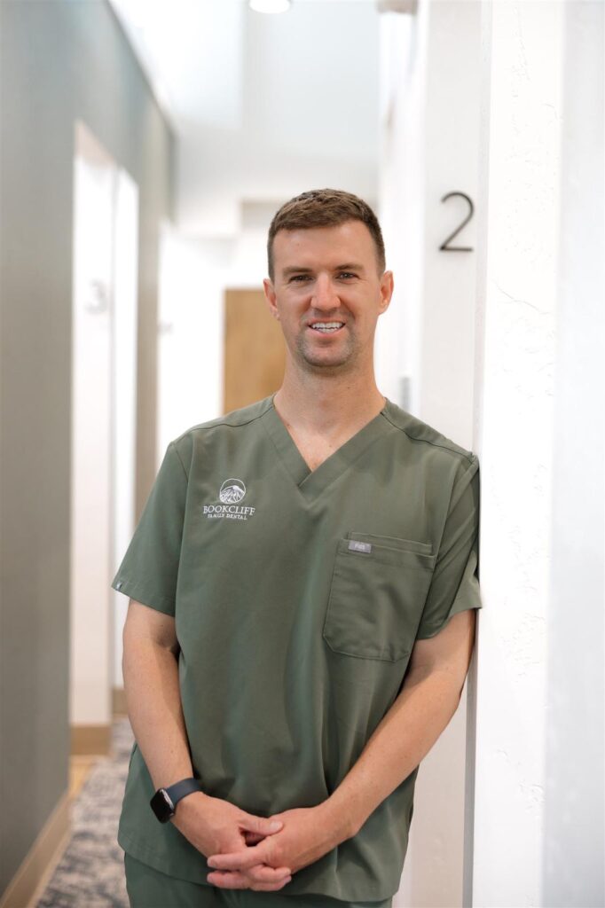Dr. Ryan of Bookcliff Family Dental in Grand Junction, CO, wearing green scrubs with clinic logo, standing in a numbered hallway designed for patient care, reflecting the clinic’s professional and welcoming atmosphere.