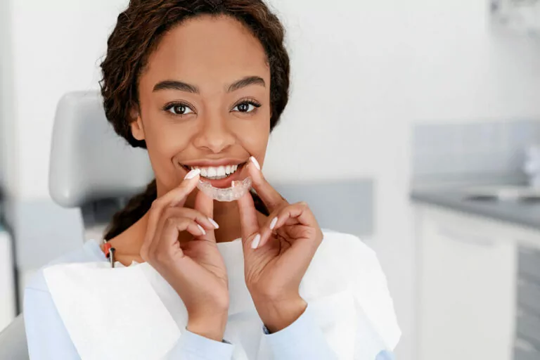 A young woman sitting in a dental chair and smiling while holding a clear dental aligner. She is wearing a dental bib and is in a clean, modern dental office. The woman is demonstrating the use of the aligner for orthodontic treatment.