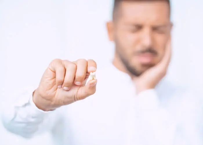 Close-up of a hand holding an extracted tooth between the thumb and index finger. In the back was the guy placing his other hand in his cheek and pain is visible to his face.