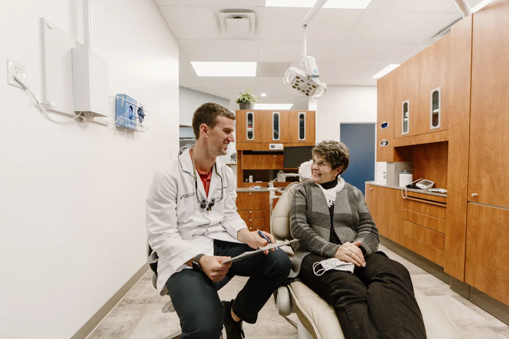 Inside a dental office, a dentist and a patient are conversing. The office has wooden cabinets, a dental light fixture, and various dental instruments.