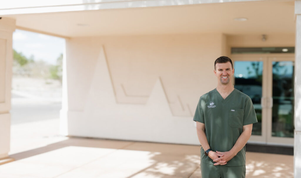 Dr. Ryan Helgerson of Bookcliff Family Dental in Grand Junction, CO, smiling in green scrubs outside the clinic’s modern entrance, showcasing a friendly, professional image