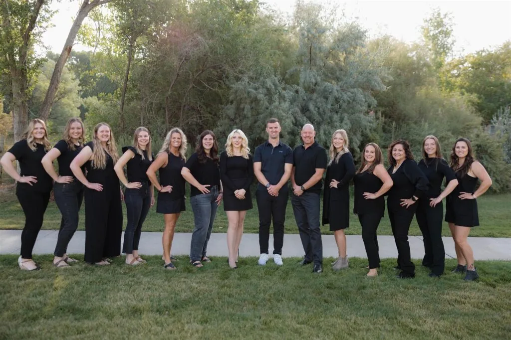 Outdoor group photo of team members from Bookcliff Family Dental, standing on a grassy area with trees and a paved path in the background. Dressed in coordinated dark attire, the team poses formally in natural daylight.