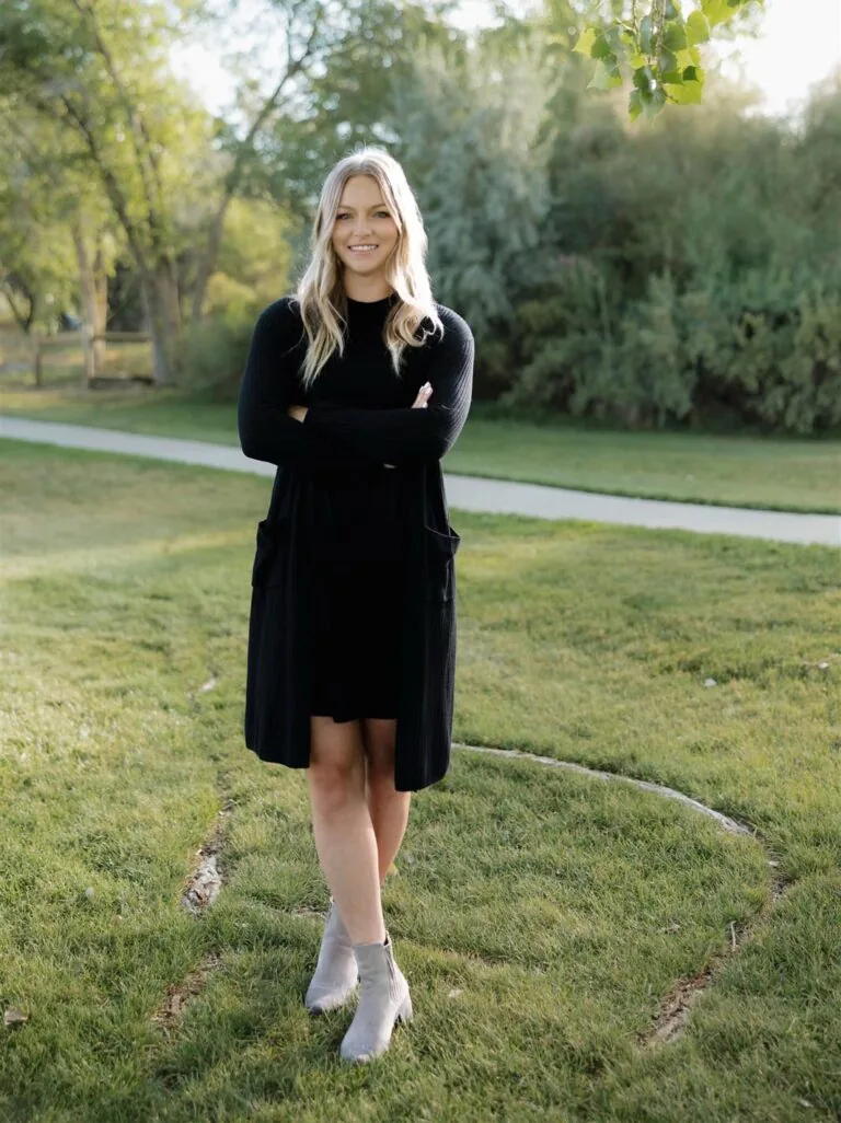 Kayla has a blonde medium length hair stands on grass in front of green foliage, wearing a black long sleeve, black skirt, and a black watch.