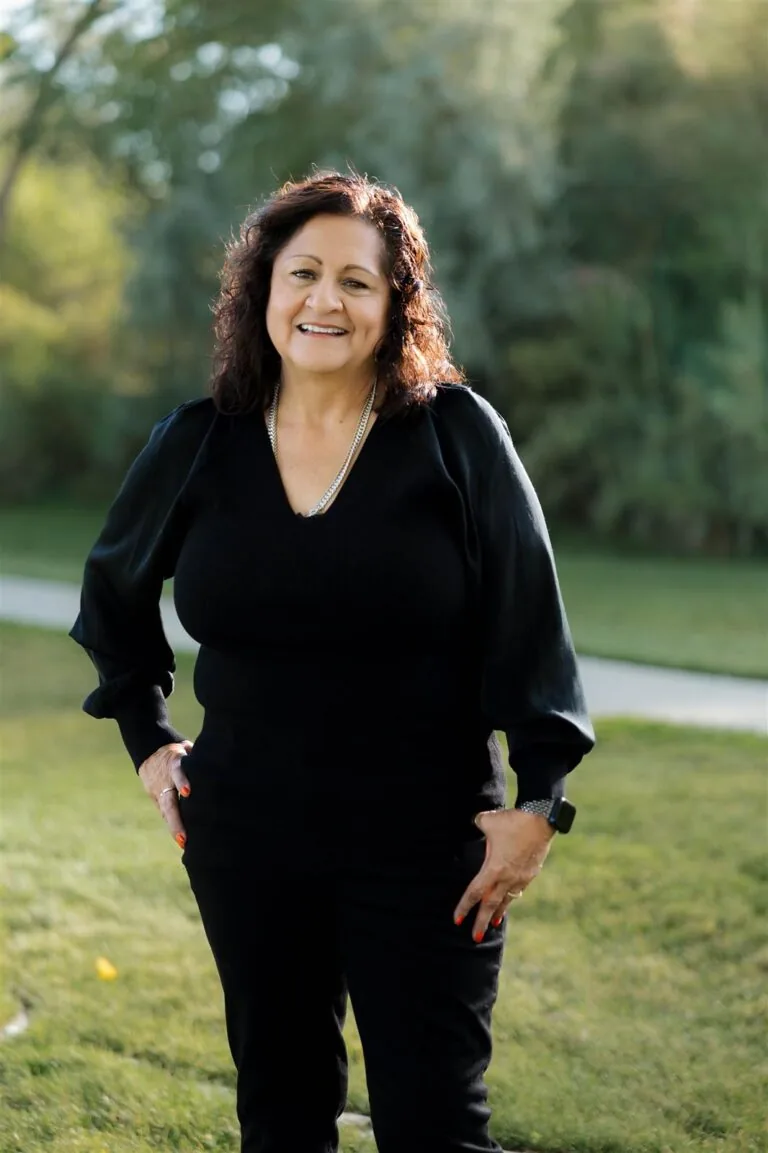 Robin has a curly shoulder-length hair stands on grass in front of green foliage, wearing a black long sleeve, dark jeans, and a black watch.