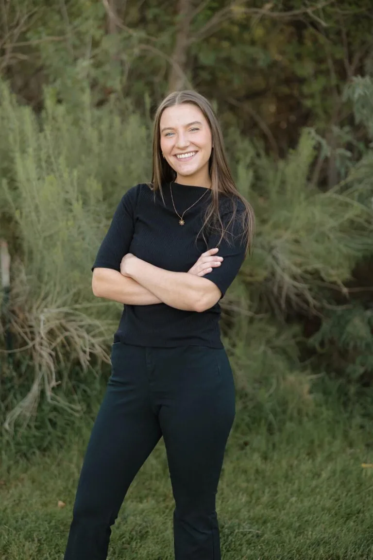 Kelsi stands on a grassy area with arms crossed, wearing black clothing and white necklace. Behind them is a landscaped space with grass and trees, suggesting a suburban or park-like setting.