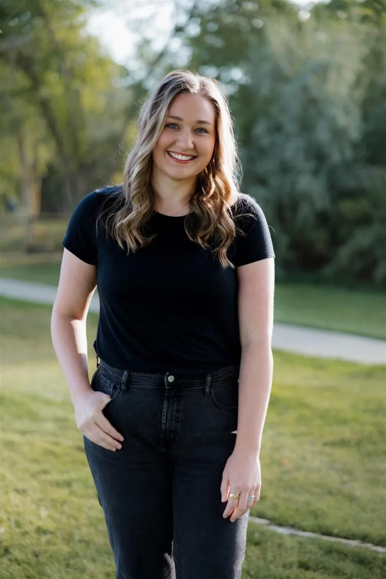 Abby has a blonde shoulder-length hair stands on grass in front of green foliage, wearing a black shirt and dark jeans.
