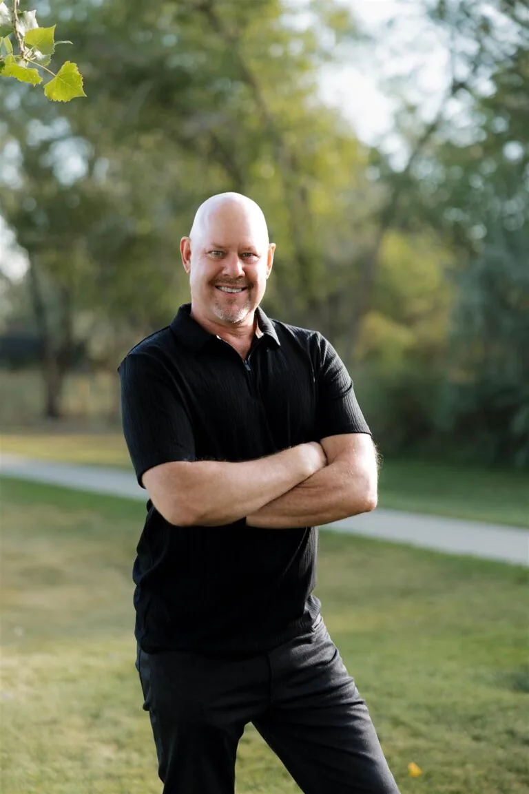 Jim stands on grass in front of green foliage, with arms crossed, wearing a black polo shirt top, and dark jeans.