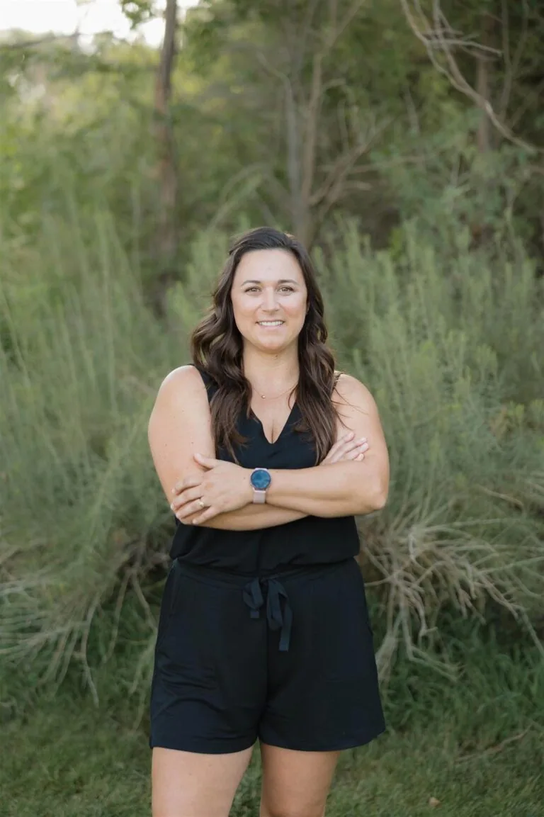 Alisha has a curly shoulder-length hair stands on grass in front of green foliage, wearing a black sleeveless , dark jeans, and a black watch .