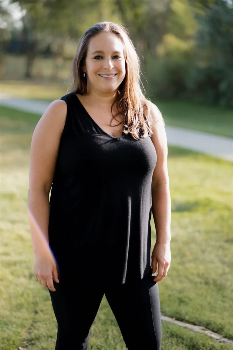 Yvette has a wavy shoulder-length hair stands on grass in front of green foliage, wearing a black sleeveless top, and dark jeans.