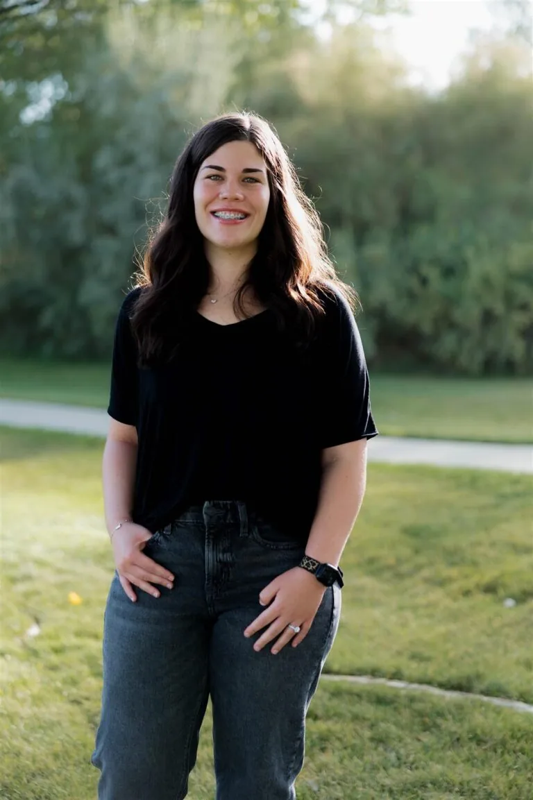 Lindsey - Dental assistant at Bookcliff Family Dental. She has a straight shoulder-length hair stands on grass in front of green foliage, wearing a black shirt , dark jeans, and a black watch.