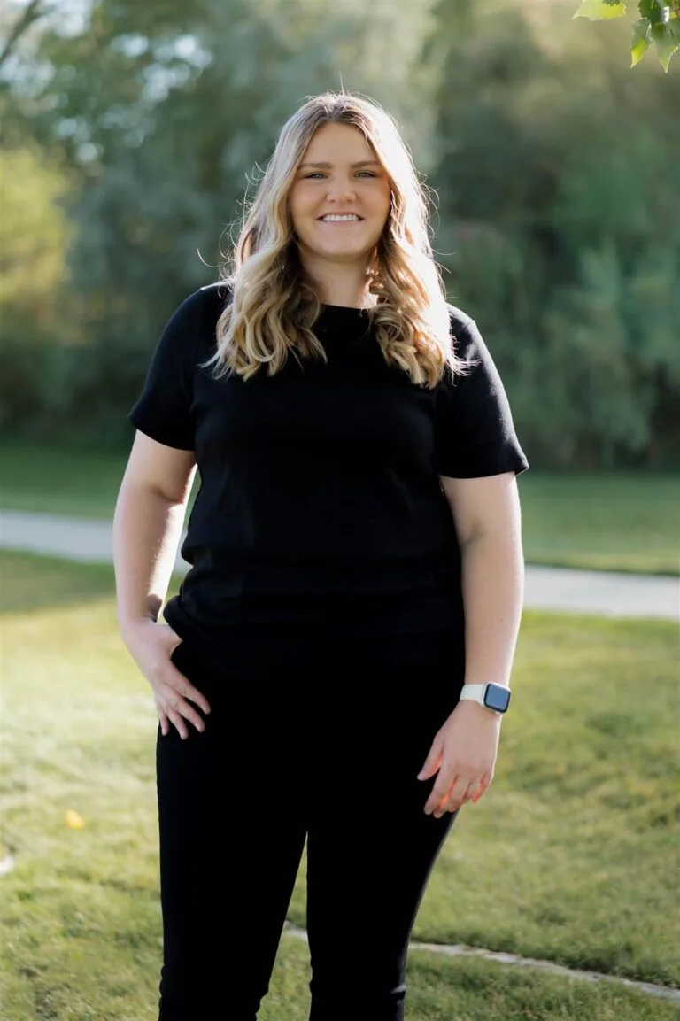 Rachel has a blonde shoulder-length hair stands on grass in front of green foliage, wearing a black shirt , dark jeans, and a black watch.