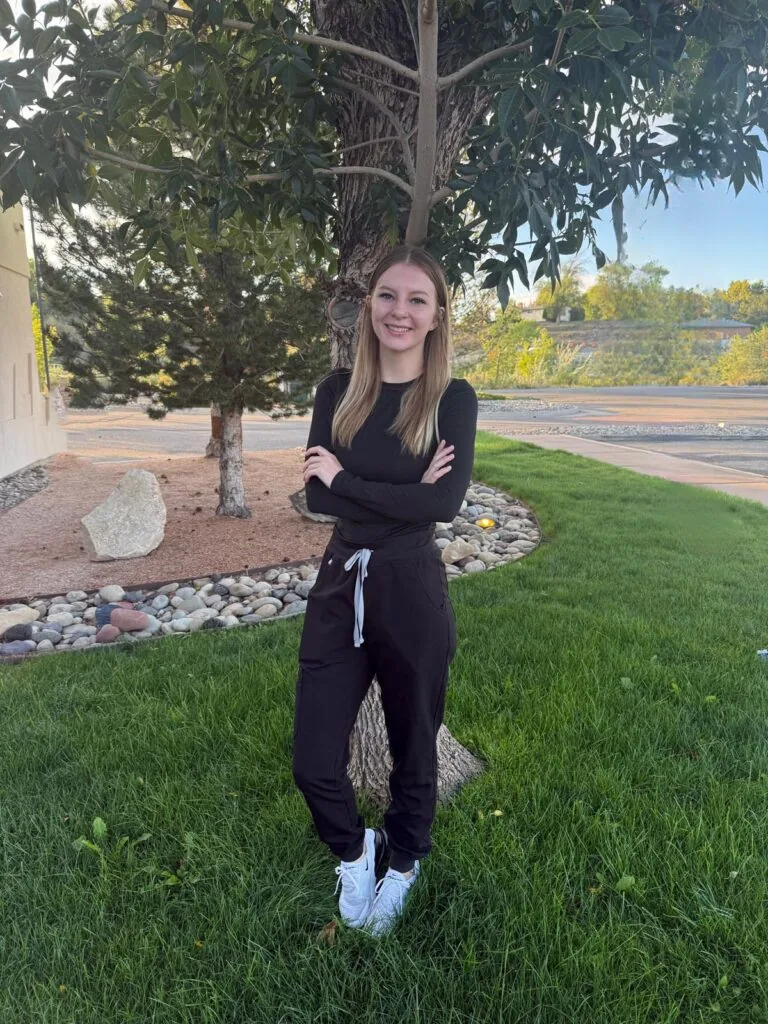 Amber Dental Assistant at Bookcliff Family Dental. Stands on a grassy area with arms crossed, wearing black clothing and white sneakers. Behind them is a landscaped space with rocks and trees, suggesting a suburban or park-like setting.