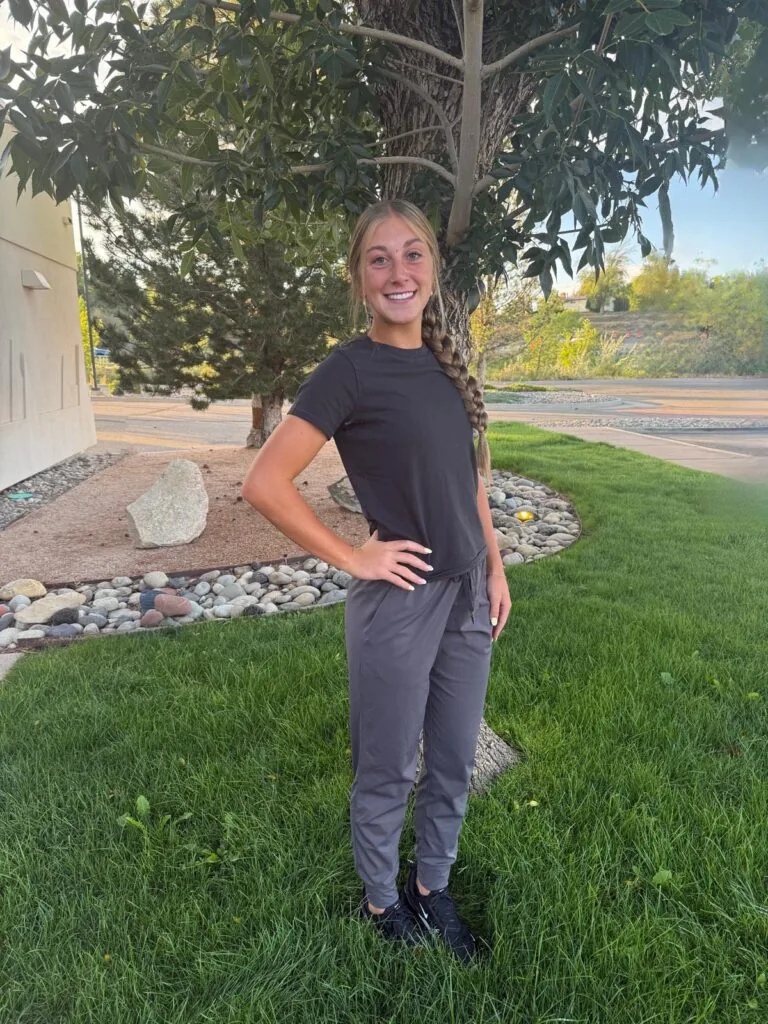 Ella Sterile Technician at Bookcliff Family Dental. Stands on a grassy area with arms crossed, wearing black clothing and white sneakers. Behind them is a landscaped space with rocks and trees, suggesting a suburban or park-like setting.